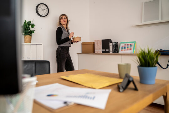 Woman standing in an office holding a lunch container with documents and a clock visible