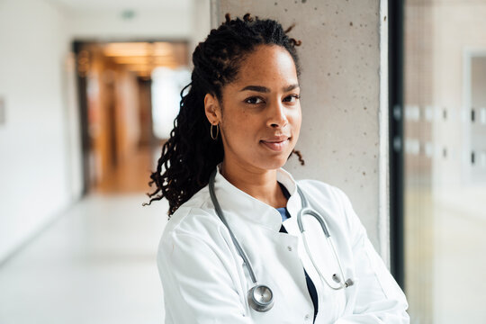 Confident young female doctor wearing lab coat leaning on column in hospital corridor