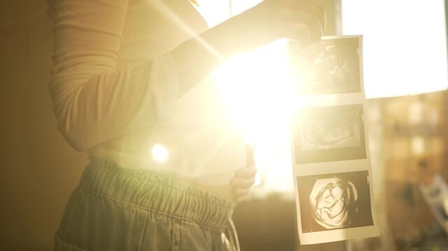 Expectant mother caressing her pregnant belly while holding ultrasound scan pictures of her unborn baby, standing by a window with warm sunlight creating a beautiful flare effect