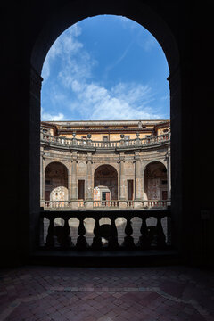 A framed view of the circular courtyard and upper loggia at Villa Farnese in Caprarola, Italy, showcasing Renaissance architecture under a blue sky.