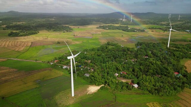 Aerial view of a rainbow arching over wind turbines and patchwork farmland landscape in Guimaras, Philippines during daytime.