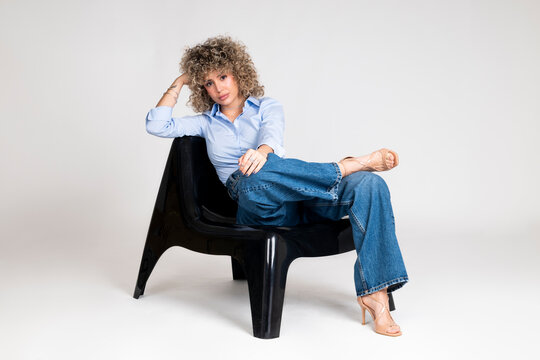 Fashionable woman with curly hair sitting confidently on a chair in a studio setting