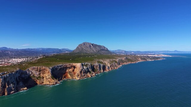 Moraira headland peninsula on the Costa Blanca, Spain, showing dramatic rocky cliffs, turquoise Mediterranean Sea and mountain backdrop under clear blue sky