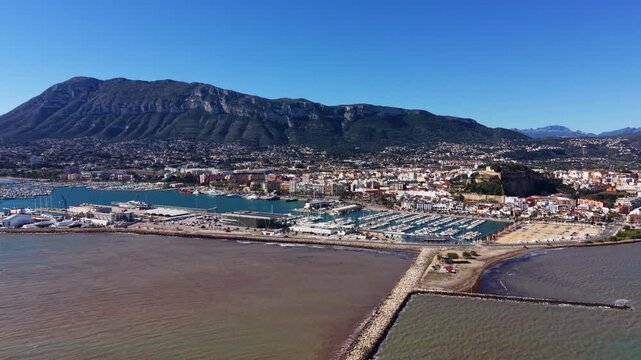 Panoramic view of Xabia marina and coastal town in Spain, with sailboats docked in harbour, white buildings, and dramatic mountain backdrop under clear blue sky