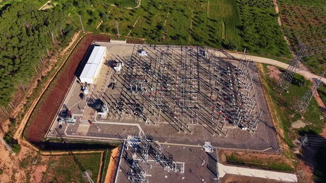Overhead view of a high voltage electrical substation surrounded by green trees and red soil, featuring transformers, switchgear, transmission lines and control building