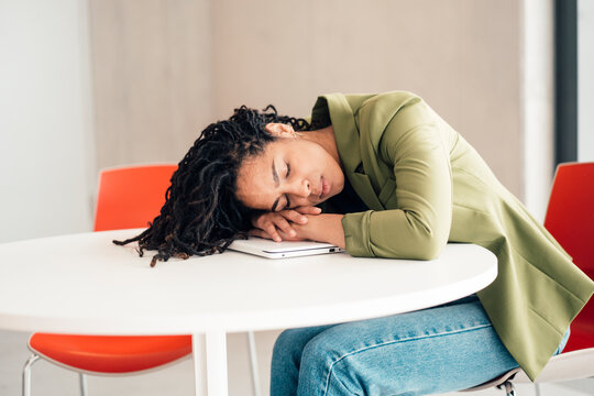 Tired young businesswoman leaning on laptop at table sleeping in office cafeteria