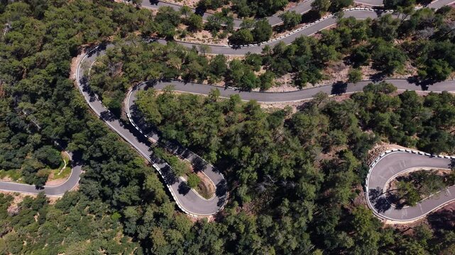 Overhead view of a winding mountain road with sharp switchbacks weaving through dense green trees on a rocky hillside in a dry landscape