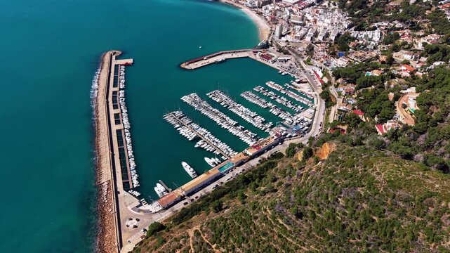 Overhead view of Xabia marina in Spain with rows of moored sailboats, turquoise Mediterranean water, breakwater pier, sandy beach and hillside town