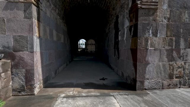 A person walks through an old stone archway with a view of the outside. The sunlight shines in, showing the texture of the stones and the path. Surroundings include grass and some structures.