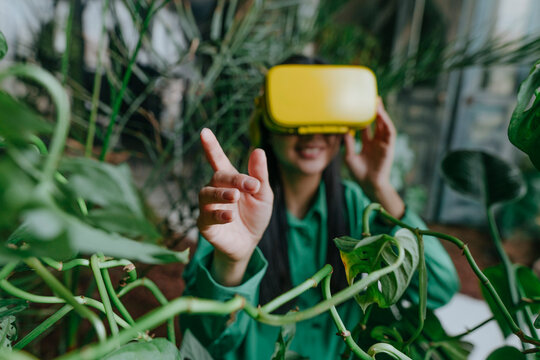 Young businesswoman wearing yellow virtual reality simulator and gesturing near plants