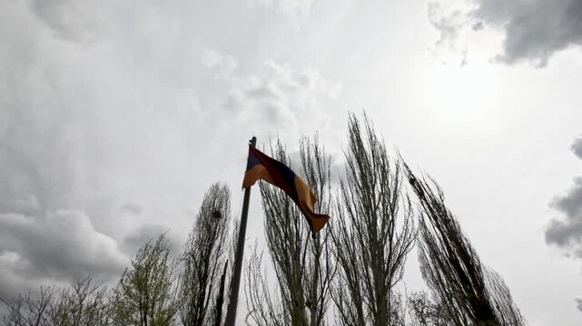 A flag is visible waving in the wind. Tall trees surround the area. The sky is cloudy with patches of light. The atmosphere shows an overcast late afternoon.
