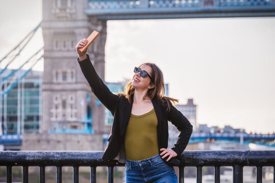 A young tourist woman is taking selfie pictures with her mobile phone in front of the Tower Bridge in London, England