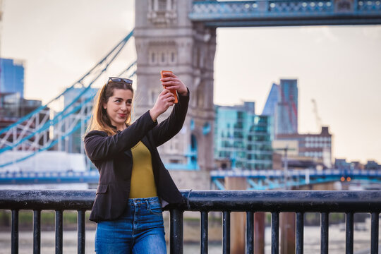 A pretty tourist woman is taking selfie pictures with her mobile phone in front of the Tower Bridge in London, England