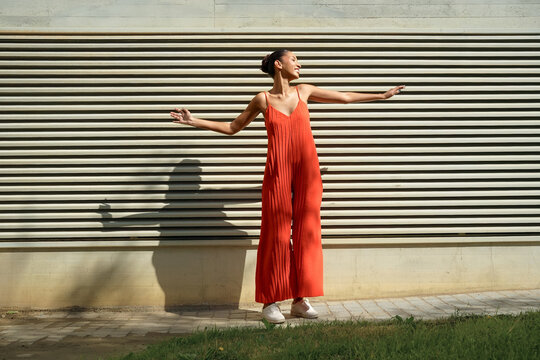 Energetic female in vivid orange clothing standing on verdant grass beside weathered metal wall