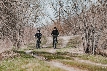 Fototapeta premium Two cyclists ride along a dirt path through a leafless winter forest. A serene and peaceful outdoor scene.