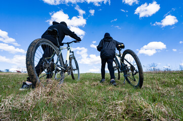Obraz premium Two cyclists walk their bikes up a grassy hill under a bright blue sky. A day of adventure awaits