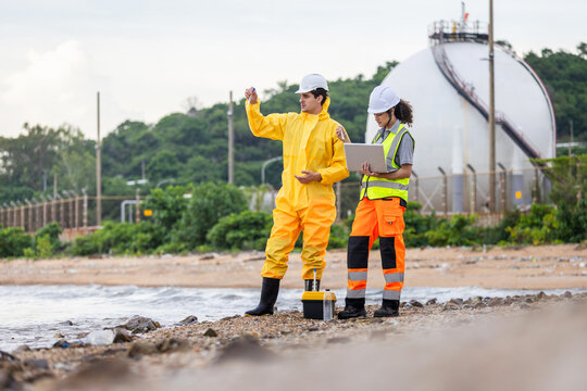 Ecologists team analyzing water pollution and quality near oil refinery or gas storage, Technical professional in protective suit and colleague with laptop monitoring industrial impact