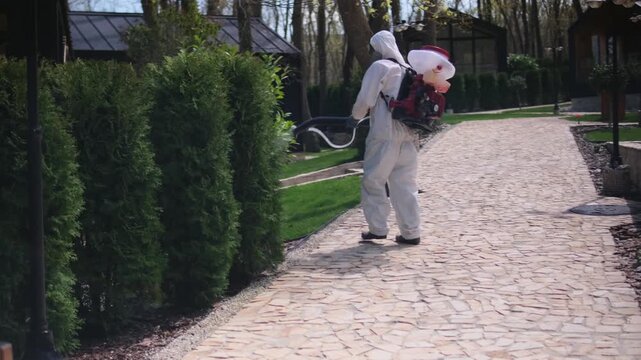 Worker in protective suit spraying garden with pesticide using backpack fogger, outdoor pest control treatment in residential yard