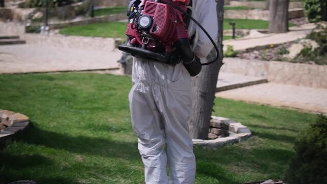 Worker in protective suit spraying garden with pesticide using backpack fogger, outdoor pest control treatment in residential yard