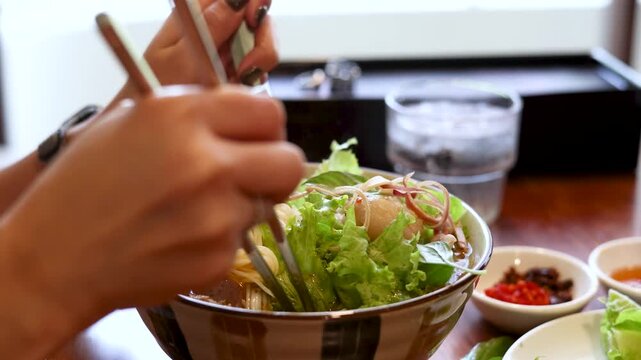 Close up shot of person hands mixing a bowl of vietnamese pho soup with chopsticks and spoon.