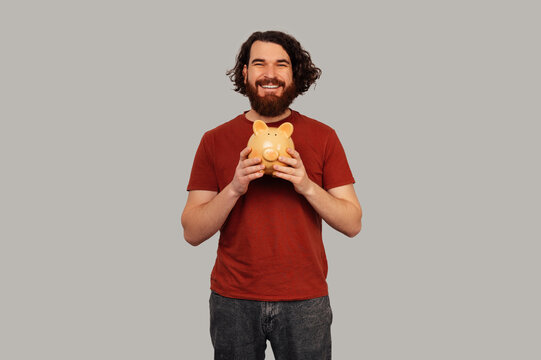 Caucasian Man Cradling Piggy Bank Close To Chest, Bearded Saver In Red Tee Conveying Thrift And Financial