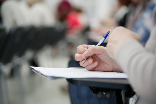 Person taking notes at a desk in a classroom or lecture hall. Close-up view of hands writing a test or essay with blurred background. Education and professional training. Photo