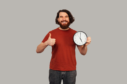 White Man Holding Clock, Thumbs Up With Broad Smile, Red Shirt And Beard, Studio Grey Background, Punctuality