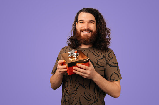 Caucasian Man Holding Wrapped Gift Box, Bearded Smiling Presenter In Patterned Shirt Against Purple Studio