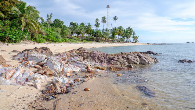 Wide landscape of Tanjung Ular beach showing coastal rocks, coconut trees, a lighthouse tower, and the blue ocean.