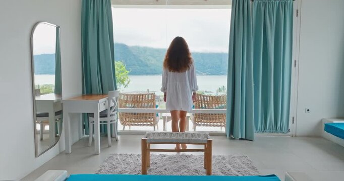 Travel to Indonesia, Bali: Woman relax on bench in hotel villa apartments, serene lake and mountain view from room balcony window, enjoy tropical vacation resort. Perfect morning scene. Back view