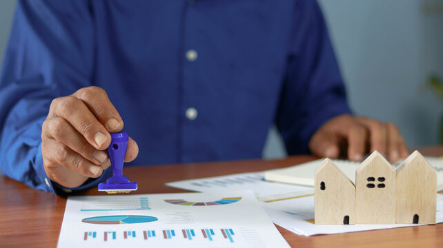 Businessman stamping real estate documents beside wooden house models, representing property approval, mortgage processing, home investment planning, legal paperwork and residential 