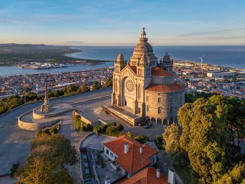 Drone photo of Santa Luzia sanctuary at sunrise above Viana do Castelo, Portugal, with a coastal panorama, terracotta rooftops, and trees framing the basilica.