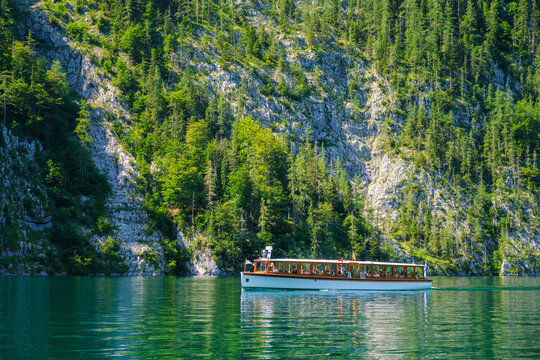 A white electric passenger boat sails on the clear emerald water of Konigsee lake by steep mountain cliffs and lush forests in Berchtesgaden National Park, Bavaria, Germany.
