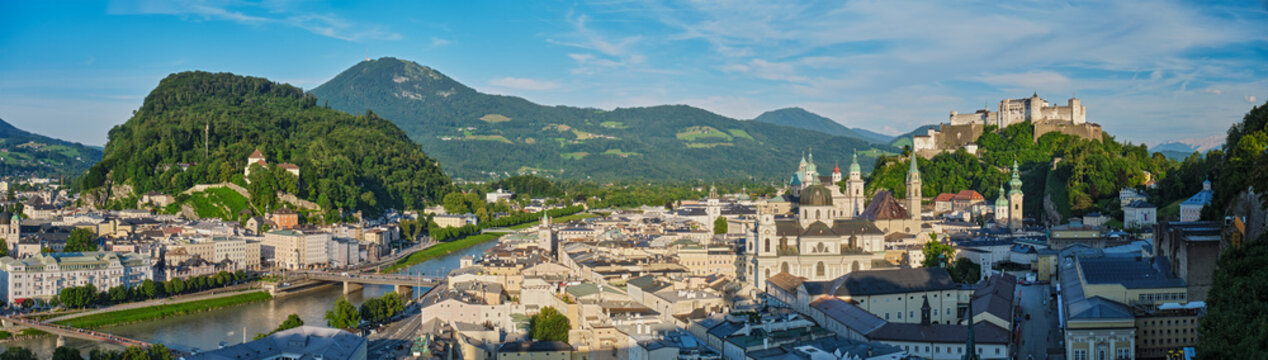 A wide panoramic aerial view of Salzburg, Austria, featuring the Salzach river, historic old town architecture, and the medieval fortress on the hill.