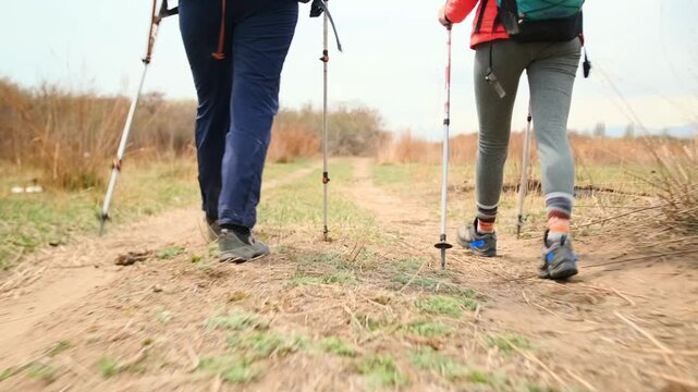 low angle rear tracking shot of two women hikers walking with trekking poles on dirt trail, focus on legs and steps, outdoor adventure, travel lifestyle, dynamic movement in nature