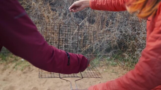Friends place sausages on metal grill grate preparing food during outdoor camping near beach, cooking simple meal in nature, enjoying travel lifestyle, relaxation and time together