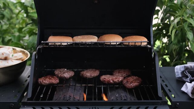 Beef burger patties grilling on barbecue with buns warming above, outdoor BBQ cooking in backyard grill