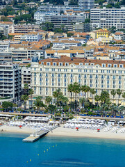 Fototapeta premium Aerial view of Cannes, France, shows Carlton Hotel facing Bay of Cannes. La Croisette lines with palms, beach clubs, sun loungers, and a pier, in crisp daylight.
