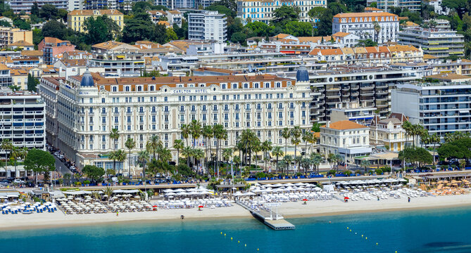 Elevated view of Cannes, France shows the Carlton Hotel on La Croisette, private beach clubs, sun loungers, a slim pier, turquoise water, and hillside apartments.