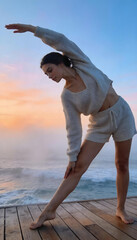 Fototapeta premium Woman doing yoga on wooden jetty at sunset. Girl wearing knitted summer resort outfit stretching by ocean. Fitness exercise at seaside, healthy lifestyle morning training concept.
