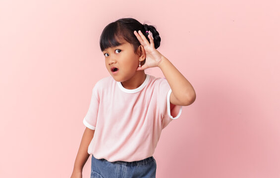 curious little asian girl eavesdropping with hand to ear, excited kid listening to secret or gossip, surprised child overhear rumors isolated on pink background.