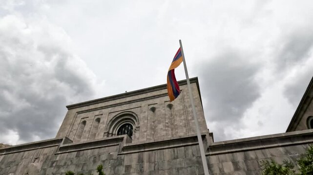 The Armenian flag flutters on a pole outside a history museum in Yerevan. It is midday and the sky is cloudy. People walk nearby as the flag moves in the wind.