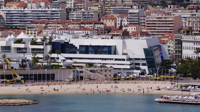 Aerial view of Cannes Croisette shows Palais des Festivals, Carlton hotel, turquoise water, sandy beach, rock jetty, white tents, flagpoles, crane, and service trucks.
