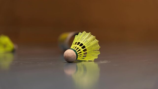 A high-visibility yellow synthetic badminton shuttlecock lying on a grey court floor. Soft lighting emphasises the texture of the feathers and cork base