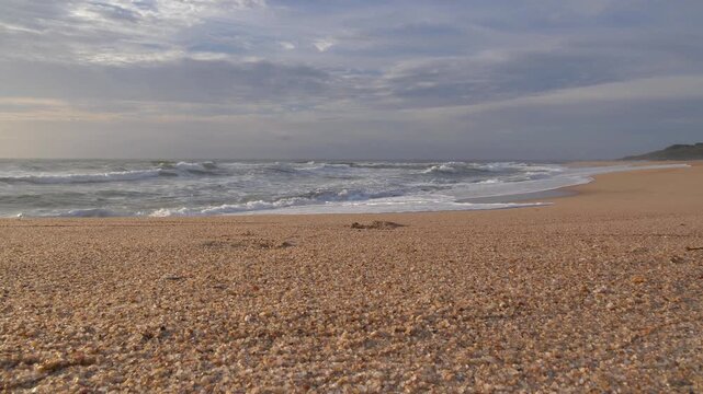 A serene coastal scene unfolds with the rhythmic motion of waves washing ashore on a vast sandy expanse, under a sky filled with soft clouds at Ramsgate, South Coast, KwaZulu-Natal.