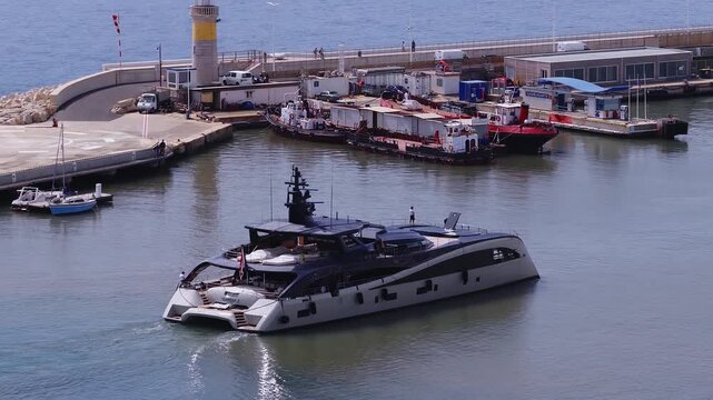 Aerial view shows a modern superyacht in Port of Cannes, France, passing CARBURANTS fuel pontoons and tug boats, crew on deck, gentle wake, bright daytime scene.