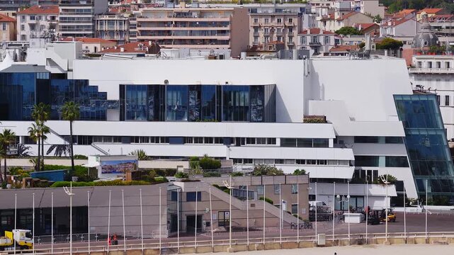 Aerial view of Palais des Festivals in Cannes, France. Trucks, a crane, and workers move along the quay as crews set up near beach, palm trees, and rooftop tents.
