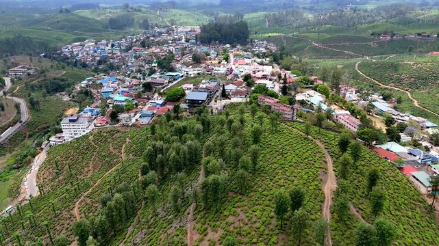 Wide aerial perspective of a summer hill town surrounded by rolling green fields and tea gardens, featuring winding roads, dense housing, and layered terrain. bright daylight enhances textures, colors