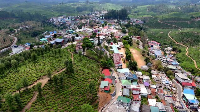 Cinematic aerial view of Valparai showcasing a vibrant hill town with colorful rooftops spreading along a ridge, surrounded by lush tea plantations and winding roads across rolling green hills.