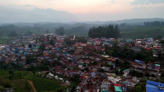 Cinematic aerial view of a hillside town at early morning, featuring soft sunrise hues, mist-covered hills, and colorful rooftops gradually illuminated by gentle light, creating a calm